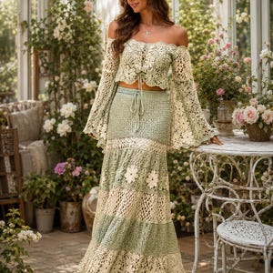 K&ouml;nnte beinhalten: Zweiteiliges H&auml;kel-Outfit in Creme und Salbeigr&uuml;n. Das schulterfreie Oberteil hat lange Glocken&auml;rmel und einen Kordelzug. Der Maxirock hat florale Details. Das Outfit wird in einem Garten getragen.