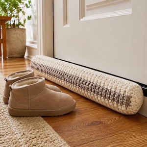 May include: A cream and brown crocheted door draft stopper rests against a white door. The cylindrical stopper is made of thick yarn with alternating stripes of cream and brown. A pair of tan suede boots with a faux fur lining sit on a beige rug.