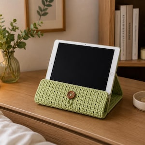 May include: A light green, crocheted tablet case with a wooden button closure. The case is holding a tablet with a black screen. The case is on a wooden surface, next to a small lamp, a vase with greenery, and a small dish.
