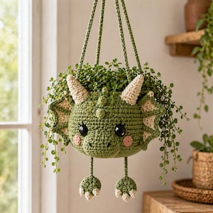 May include: A green crocheted dragon-shaped hanging planter filled with greenery. The planter features large black eyes, beige horns, and pink cheeks. The dragon's feet dangle below. The planter is suspended by green rope.