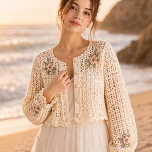 Puede incluir: Rebeca de crochet color crema con detalles florales. El diseño calado presenta aplicaciones de flores rosas y verdes en las mangas y el corpiño. La rebeca tiene un dobladillo festoneado y botones perlados.