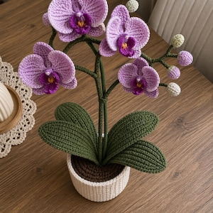 Può includere: Una pianta di orchidea all'uncinetto fatta a mano in un vaso bianco. L'orchidea presenta grandi fiori viola-lavanda con centri viola scuro e gialli. Foglie e steli verdi completano il design. Il vaso è realizzato con filato color crema.