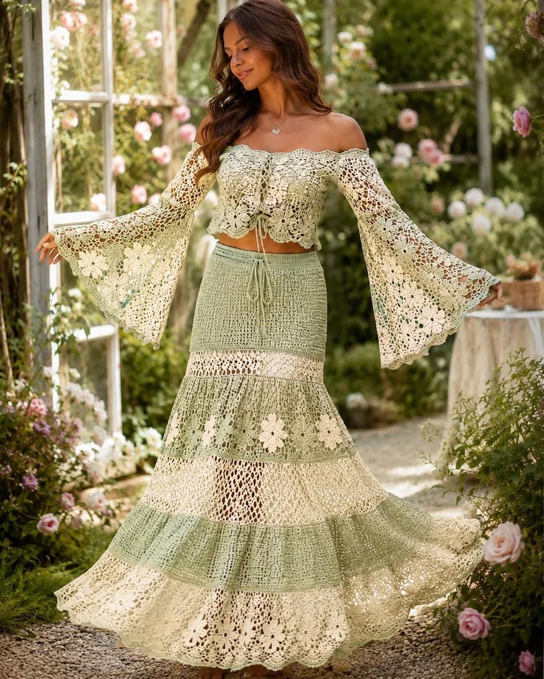 K&ouml;nnte beinhalten: Zweiteiliges H&auml;kel-Outfit in Gr&uuml;n- und Cremet&ouml;nen. Das schulterfreie Oberteil hat lange, ausgestellte &Auml;rmel. Der Rock ist ein Stufen-Design mit floralen Akzenten. Das Outfit wird im Freien getragen, umgeben von Gr&uuml;n und Blumen.