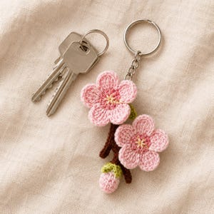 May include: A crochet keychain featuring two pink cherry blossom flowers and a small bud attached to a brown stem, connected to a silver metal split ring. Two silver metal keys are attached to the ring, which has a chain link connector.