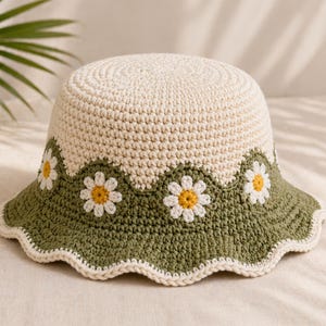 May include: A cream-colored crochet bucket hat with a green scalloped brim. The brim is decorated with white daisies with yellow centers. The hat is a stylish accessory for sun protection.