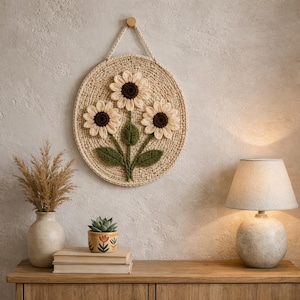 May include: A beige, oval-shaped wall hanging featuring three crocheted sunflowers with brown centers and green leaves. The sunflowers are attached to a woven background. The wall hanging is suspended by a beige cord and a wooden peg.