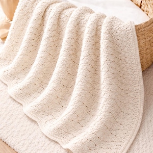 May include: Cream-colored baby blanket with a textured, woven design. The blanket is draped over a woven basket with a white interior. The blanket is made of soft material and is placed on a matching rug.