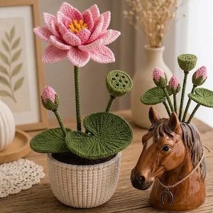 以下が含まれることがあります： 白い鉢に入った手作りのかぎ針編みの蓮の花、緑の葉とつぼみ。その隣には、蹄鉄のペンダントが付いた茶色のセラミックの馬の頭の形をした花瓶があります。蓮の花はピンク色で、中心は黄色です。