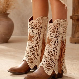 May include: Brown leather cowboy boots with cream crochet boot cuffs. The cuffs have a star design and fringe. The boots have a pointed toe and a low heel, suitable for a bohemian or country style.