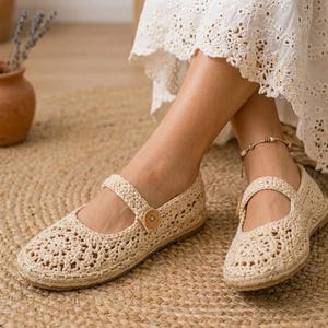 Könnte beinhalten: Beige Häkel-Mary-Jane-Ballerinas mit Riemen und Holzknopfverschluss. Die Schuhe haben eine gewebte Textur und eine runde Spitze. Die Ballerinas werden auf einem gewebten Jute-Teppich gezeigt.