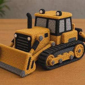 Op de afbeelding: Een handgemaakte gehaakte bulldozer in geel, grijs en zwart. Het speelgoed heeft een voorblad, rupsbanden en een cabine met ramen. De bulldozer is een gedetailleerd, zacht speelgoed, voor kinderen.