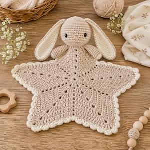 Könnte beinhalten: Ein handgehäkeltes Häschen-Kuscheltuch in Beige mit einer sternförmigen Decke. Das Häschen hat lange Ohren mit cremefarbenen Innenseiten und schwarzen Augen. Die Decke hat einen gewellten Rand und ein detailliertes Lochmuster. Ein sternförmiger Beißring aus Holz ist sichtbar.
