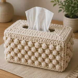 May include: A rectangular, cream-colored tissue box cover made of crocheted yarn. The cover features a textured, bobble stitch design on the sides and a hole on top for tissue dispensing. White tissues are visible. The box is on a neutral-toned surface.