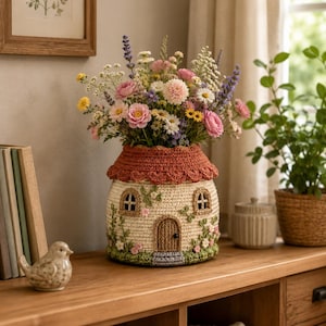 Pode incluir: Um vaso artesanal em forma de casa de crochê, cheio de um vibrante buquê de flores. O vaso apresenta um telhado vermelho, pequenas janelas e uma porta, com enfeites florais. O vaso está sobre uma superfície de madeira.