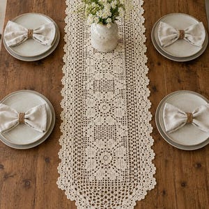 May include: A cream-colored, crocheted table runner with a floral design, set on a wooden table. The table is set with four plates, each with a linen napkin tied with twine. A vase of white flowers sits in the center.