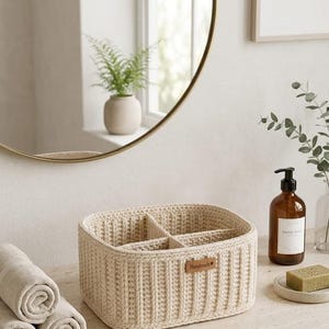 May include: A cream-colored, rectangular crochet basket with four compartments, labeled "Handmade." Rolled towels and a soap dispenser are nearby. A round mirror and a framed print are in the background. The basket is designed for bathroom organization.