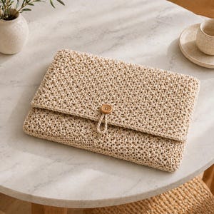 Puede incluir: Una funda beige para portátil de ganchillo con cierre de solapa y un botón de madera. La funda es rectangular y tiene un aspecto texturizado. El botón es redondo y marrón. La funda está sobre una mesa de mármol.