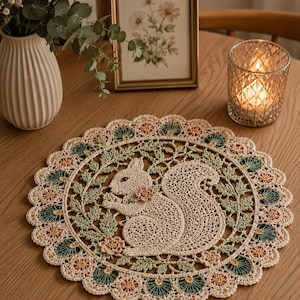 May include: A cream-coloured, crocheted doily with a squirrel design. The squirrel is surrounded by green leaves and small flowers in shades of pink, green, and cream. The doily has a scalloped edge.