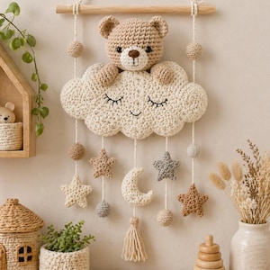 May include: Crocheted nursery wall hanging featuring a tan teddy bear peeking over a cream-colored cloud with closed eyes. Hanging below are stars, a crescent moon, and a tassel. The wall hanging is suspended from a wooden dowel.