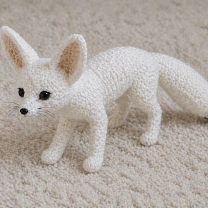 Può includere: Una volpe Fennec bianca fatta a mano all'uncinetto con grandi orecchie e una coda con la punta nera. La volpe ha occhi neri e baffi. L'animale artigianale è posizionato su una superficie chiara e strutturata.