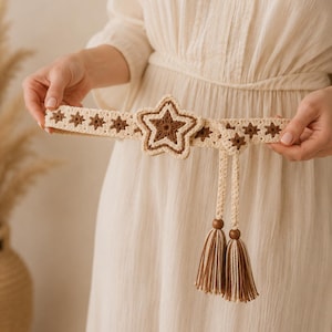 May include: A cream-coloured crochet belt with a large star-shaped buckle and smaller star accents. The belt features brown and cream tassels with wooden beads. The belt is approximately 7.6 cm wide.