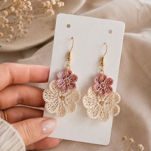 Puede incluir: Pendientes de flores de ganchillo hechos a mano. Los pendientes presentan una base de flor color crema con una flor malva más pequeña en la parte superior. Se utilizan ganchos dorados y pequeñas cuentas doradas en el diseño. Los pendientes se muestran en una tarjeta blanca.