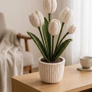Op de afbeelding: Een gehaakt arrangement van witte tulpen in een witte pot. De handgemaakte bloemen hebben groene stelen en bladeren, en de pot staat op een houten tafel. De opstelling omvat vijf tulpen en een gebreide pot.
