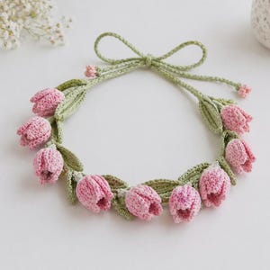Könnte beinhalten: Ein handgefertigter gehäkelter Blumenkranz mit rosa tulpenförmigen Blüten und grünen Blättern. Der Kranz ist mit einer grünen Schleife gebunden und eignet sich für dekorative Zwecke.