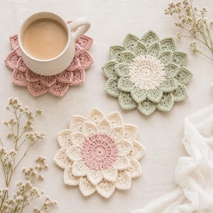 Pode incluir: Três bases para copos em croché com formato de flor e pétalas em camadas. Uma base rosa seco está sob uma caneca de cerâmica, uma é verde sálvia com centro creme e uma é creme com o centro rosa seco.