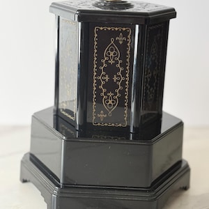 May include: A black hexagonal container with a tiered base and a lid featuring a small central knob. The vertical panels of the upper section are decorated with intricate gold-colored line patterns, while the base remains plain and glossy black.