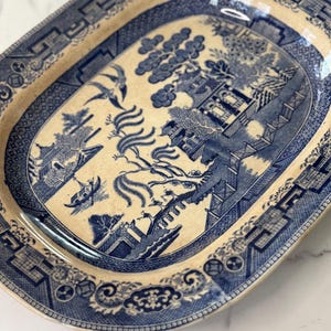 Peut inclure: Un plat de service ovale en céramique avec un motif willow bleu et crème. Le décor représente un paysage avec une pagode, un pont, un saule et deux oiseaux en vol. La bordure est ornée de motifs géométriques et floraux détaillés.