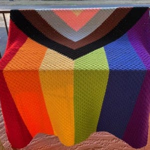 May include: A colorful, handmade crochet blanket with a geometric design. The blanket features a vibrant rainbow of colors, including red, orange, yellow, green, blue, and purple. The blanket is displayed outdoors, draped over a railing.
