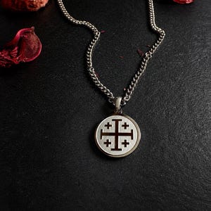 May include: A silver-toned metal necklace featuring a circular pendant with a Jerusalem cross emblem. The pendant has a textured, light-colored center with a dark cross design and is suspended from a matching silver-toned curb chain link necklace.