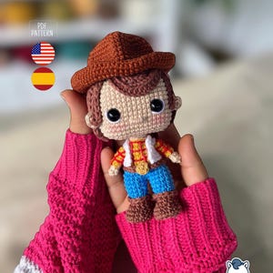 May include: A handmade crochet doll of a cowboy character, featuring a brown hat, hair, and boots, with a yellow shirt, white vest, and blue jeans. The doll is held in pink knitted gloves. The image includes a "PDF PATTERN" label and flags.