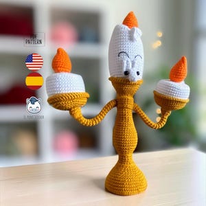 May include: A crochet amigurumi figure shaped like a candelabra with a yellow base and arms, white candle bodies, and orange flames. The central candle features a white face with black embroidered eyes and eyebrows. Text reads PDF PATTERN.