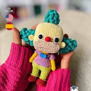 Könnte beinhalten: Handgefertigte gehäkelte Clown-Puppe mit gelbem Kopf, türkisfarbenem Haar und großen schwarzen Augen. Der Clown trägt ein lila Hemd, grüne Hose und rote Schuhe. Die Puppe wird in den Händen einer Person gehalten, mit einem rosa Pullover im Hintergrund.