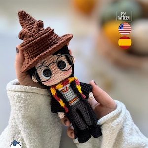 Puede incluir: Muñeca de ganchillo hecha a mano de un personaje con un Sombrero Seleccionador marrón, gafas redondas y una bufanda roja y amarilla. La muñeca viste una túnica negra y pantalones grises. La imagen incluye el texto "PDF PATTERN" y las banderas de Estados Unidos y España.