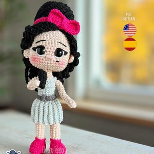 Peut inclure: Poupée au crochet faite à la main avec des cheveux noirs, un nœud rose et une robe bleu clair. La poupée a des chaussures roses et des joues roses. La poupée tient une mèche de ses cheveux. Le patron est en PDF.