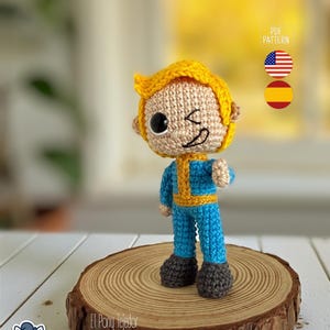 Puede incluir: Figura de ganchillo de un personaje con pelo rubio, guiñando un ojo, con un mono azul con detalles amarillos y botas grises. La figura está sobre una rodaja de madera. La imagen incluye el texto "PDF PATTERN" y banderas de EE. UU. y España.