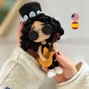 Può includere: Bambola all'uncinetto fatta a mano di un musicista, con un cappello a cilindro nero con accenti blu, occhiali da sole rotondi e che tiene una chitarra gialla. La bambola ha capelli ricci neri e scarpe bianche. Il testo "PDF PATTERN" è visibile.
