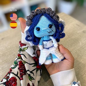 May include: A handmade crochet doll with a blue face, dark blue hair, and a white dress. The doll has a stitched mouth and black eyes. The doll is held in a person's hands. The doll is a PDF pattern.