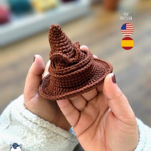 May include: A small, brown crocheted hat, resembling a wizard's sorting hat, is held in a person's hands. The hat is made of a textured yarn and features a pointed top and a wide brim. The image includes the text "PDF PATTERN".