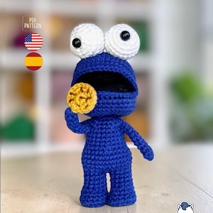 Puede incluir: Muñeco de ganchillo azul de Cookie Monster con ojos blancos grandes y una galleta amarilla. El muñeco tiene la boca abierta y sostiene la galleta. La imagen incluye el texto "PDF PATTERN" y las banderas estadounidense y española.