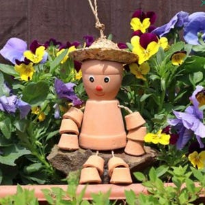 May include: A terracotta pot figure with a straw hat, arms, and legs made of smaller pots. The figure has painted eyes and a red nose, set amidst vibrant purple, yellow, and blue pansies. A charming garden ornament.