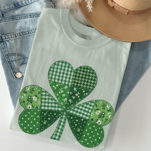 Può includere: Una maglietta verde chiaro con un grande motivo a trifoglio realizzato con vari tessuti verdi fantasia. La maglietta è appoggiata su jeans azzurri, con una collana d'oro e un cappello marrone sullo sfondo. Un tema del giorno di San Patrizio.