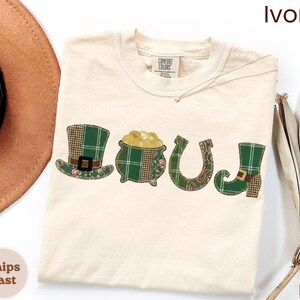 May include: Ivory-colored t-shirt with the word "LUCK" spelled out in patchwork designs. The letters feature a green plaid top hat, a pot of gold, a horseshoe, and a green boot with a buckle. The shirt has a Comfort Colors tag.
