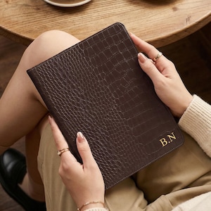 May include: A dark brown leather journal with a crocodile skin effect is held. The journal has gold initials "B.N". A cup of coffee and a pastry are on a wooden table in the background. The journal is likely A5 size, approximately 21 cm x 15 cm.