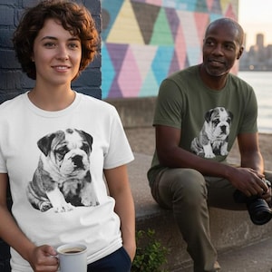 May include: Two people wearing t-shirts with a black and white bulldog puppy graphic. One shirt is white, the other is olive green. The background includes a brick wall, a colorful mural, and a waterfront scene.