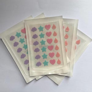 May include: Four sealed packages of acne patches. Each package contains patches in the shapes of stars, hearts, clouds, and teardrops in pastel colors of pink, purple, and teal. The packages are arranged on a white surface.