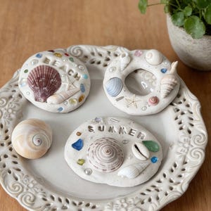 May include: A white ceramic plate with a decorative edge holds three small, round, beach-themed ornaments. The ornaments are decorated with seashells, colorful glass beads, and the words "BEACH" and "SUMMER". A small potted plant sits nearby.